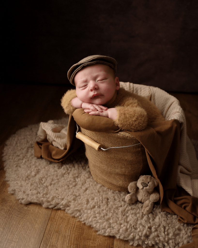 A sleeping newborn baby dressed in autumnal colours with a brown flat peak cap. Positioned in a basket with blankets draped over