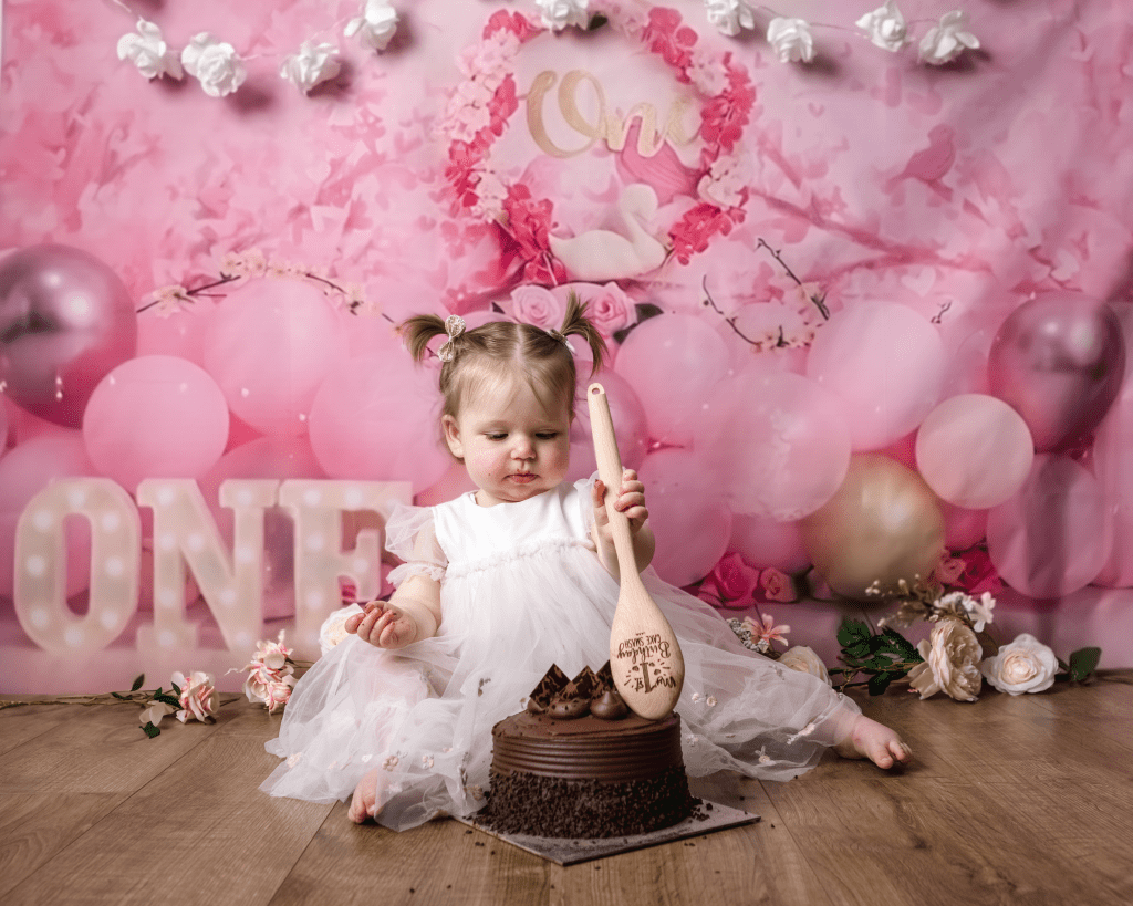 a one year old little girl in a pink dress sitting on the floor in front of a cake. She is holding a wooden spoon about to dig in.