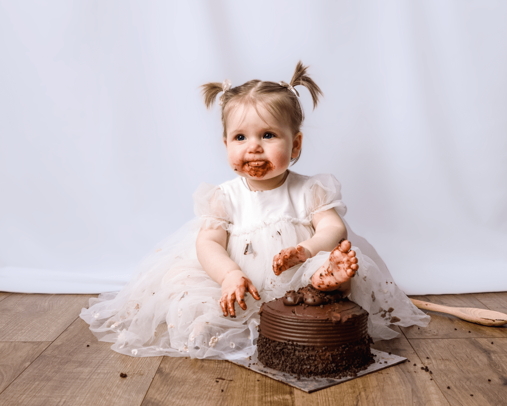 a little girl with the tongue sticking out had chocolate cake all over her hands and face. Her left foot is on the cake.