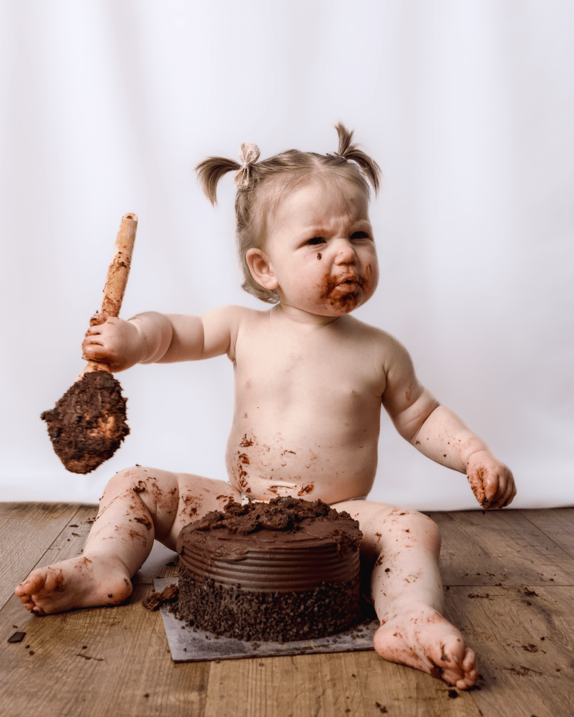 a little girl sits proudly in front of a cake, holding a wooden spoon, she is covered in chocolate cake after smashing it up
