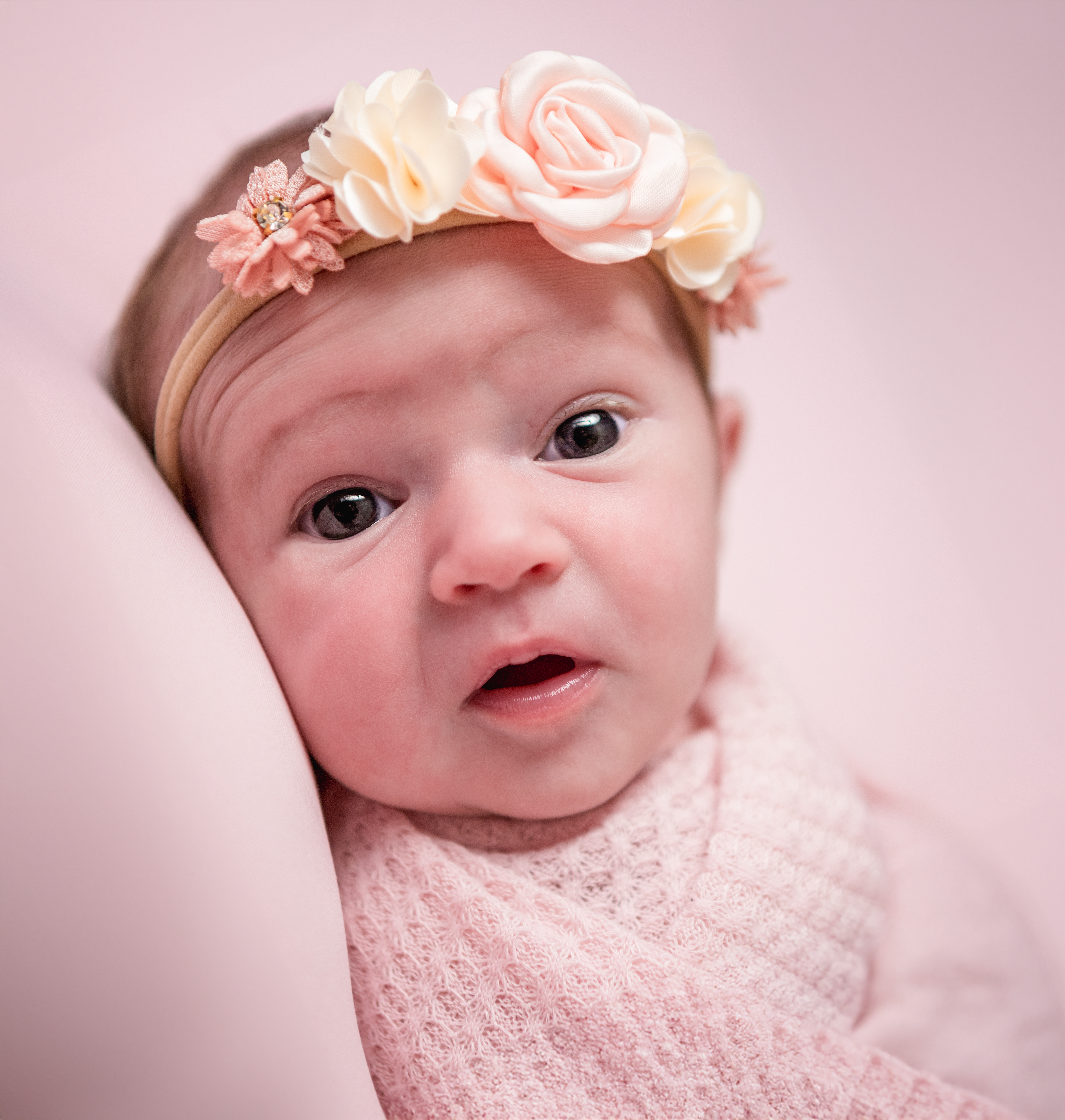 newborn baby with wide eyes looking into the camera. she is wearing a pink headband