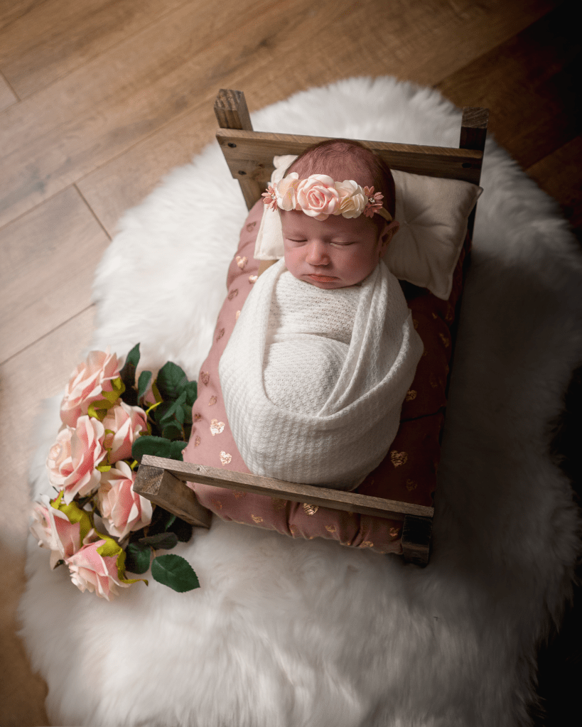 a newborn baby, swaddled, sleeping on a tiny bed with a floral headband